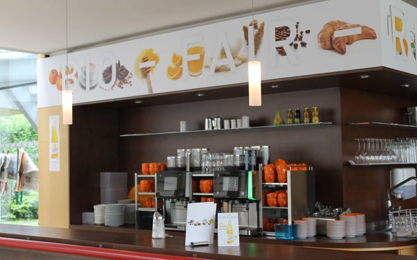 Bar im Haus der Begegnung. Hinter der Theke sind zwei Kaffeemaschinen, Tassen, Gläser und Teller. Über der Bar steht "Bio - Fair - Regional".