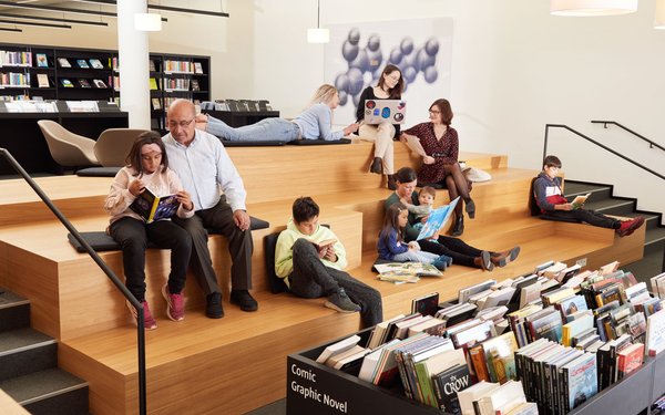 Ein Raum in der Stadtbibliothek mit breiten Holzstufen, auf denen Menschen verschiedener Altersgruppen sitzen und lesen. Vorne und hinten sieht man Bücherregale.