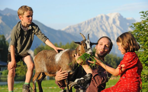 Ein Mann, ein Junge und ein Mädchen kümmern sich um eine Ziege und füttern sie mit Gras vor einem ländlichen Hintergrund.