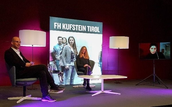 Podium mit einem Mann, einer Frau und einem Bildschirm, auf dem eine Frau zu sehen ist. Im Hintergrund stilvolle Beleuchtung und ein RollUp der FH Kufstein.