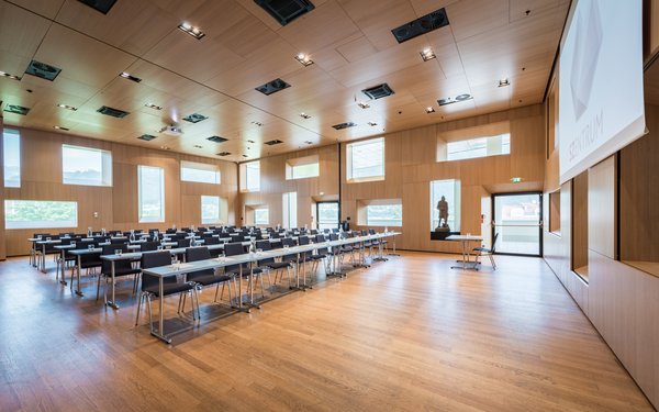 Großer, heller Saal in Holzoptik mit Reihenbestuhlung, Tischen und Leinwand mit Beamer.