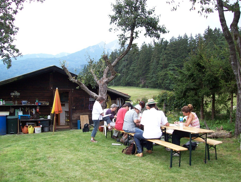 Vor einer Hütte, umgeben von einer Wise und einem Wald im Hintergrund, sitzen mehrere Menschen an einem Tisch und nehmen an einem Kurs teil.