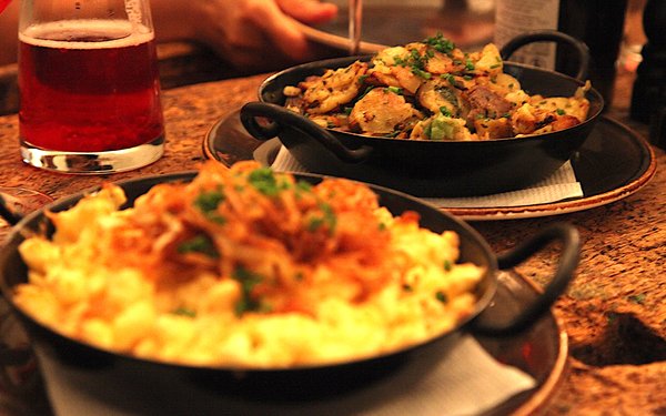 Käsespätzle und ein Gröstl in Pfandln, daneben ein Krug Bier.