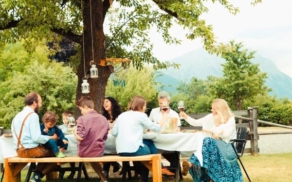Festlich gekleidete Menschen essen an einer Gartentafel unter einem Baum im Grünen und stoßen mit Wein an.