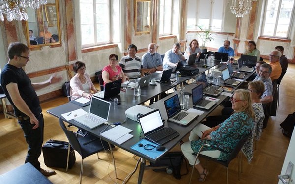 Im barocken Spiegelsaal findet eine Arbeitsveranstaltung statt. In der Mitte des Raumes steht ein großer, schwarzer Besprechungstisch. Rundherum sitzen Menschen mit Laptops, während ein stehender Mann am Anfang des Tisches die Veranstaltung leitet.